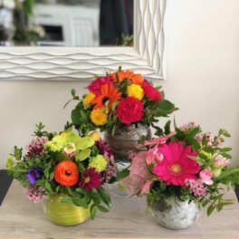 Three small mixed flower arrangements with bright pink, orange, and yellow blooms in round containers.