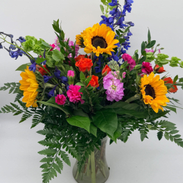 Vibrant mixed bouquet with sunflowers, pink and orange blooms, and blue delphinium in a clear glass vase.
