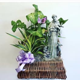 Potted plant arrangement with a saint statue and purple ribbon in a wicker basket