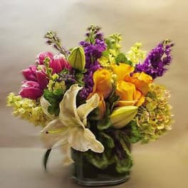 Mixed bouquet of yellow roses, white lilies, pink tulips, and purple flowers in a glass vase