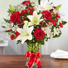Arrangement of red roses, red carnations, and white lilies in a clear glass vase with a red ribbon bow