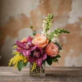 Peach roses, purple orchids, yellow accents, and white snapdragons in a clear glass cube vase