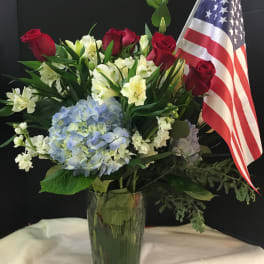 Red roses and white lilies with blue hydrangea in a glass vase beside an American flag