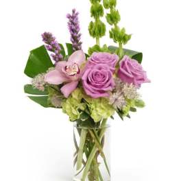 Tall arrangement of lavender roses, green hydrangea, Bells of Ireland and a pink orchid in a clear glass vase