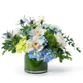 Low glass vase arrangement with blue hydrangeas, white roses, orchids, and green accents