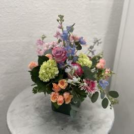 Pastel arrangement of pink and peach roses, green hydrangea and blue blooms in a green square vase