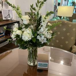 White flower arrangement with roses and snapdragons in a clear glass vase beside a small boxed gift.