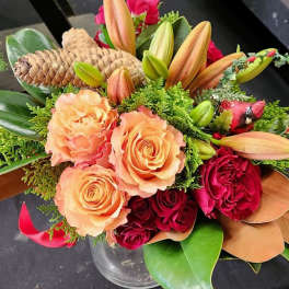 Peach and red roses with orange lilies, pinecones, and a red cardinal pick in a clear vase.