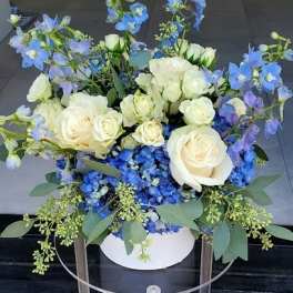 White roses and blue flowers arranged in a round white box on a clear stand