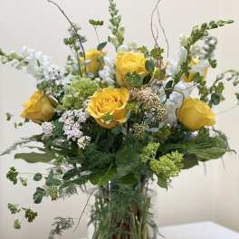 Tall arrangement of yellow roses, white snapdragons, and green accents in a clear glass vase