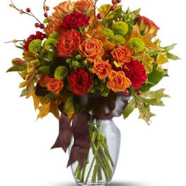 Autumn arrangement of orange roses, red carnations, green mums and fall leaves in a clear glass vase