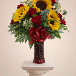 Tall arrangement of yellow sunflowers and red roses in a red glass vase with a red bow.