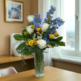 Tall arrangement of white roses, blue delphinium, and yellow blooms in a clear glass vase on a wooden table