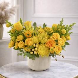 Low yellow flower arrangement with roses, protea, and mums in a white ceramic pot on a marble table