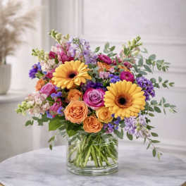 Bright mixed bouquet of yellow gerbera daisies and roses in a clear glass vase on a round table.