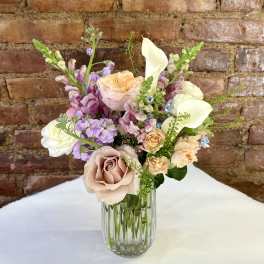 Pastel bouquet of roses, calla lilies, and purple blooms in a glass vase