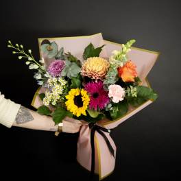 Handheld bouquet of orange, pink, and burgundy flowers wrapped in teal paper