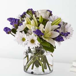 White lilies and daisies with purple filler in a clear glass vase