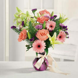 Mixed bouquet of pink and coral flowers in a purple glass vase with a ribbon