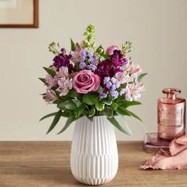 Mixed bouquet of pink roses and purple flowers in a white textured ceramic vase on a wooden table