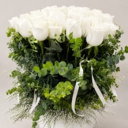 White roses arranged in a round white vase with lush greenery and ribbon accents