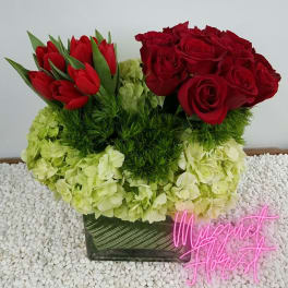 Red roses and tulips arranged with pale green hydrangeas in a square vase.