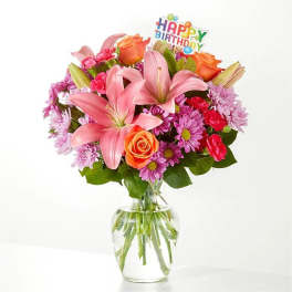 Mixed bouquet of pink lilies, orange roses, and purple mums in a clear glass vase with a Happy Birthday pick.