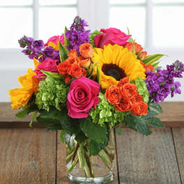 Bright mixed bouquet of roses, sunflowers, and purple blooms in a glass vase