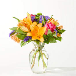 Bright mixed bouquet of lilies, roses, and purple filler flowers in a clear glass vase