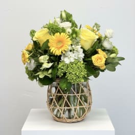 Yellow and white flower arrangement in a glass vase with wicker wrap