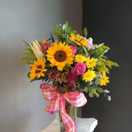 Mixed bouquet of sunflowers, roses, and lilies in a glass vase with a pink plaid ribbon.