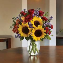 Bouquet of sunflowers, red roses, and purple flowers in a clear glass vase