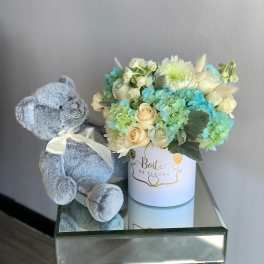 Blue teddy bear beside a pastel flower arrangement in a white box