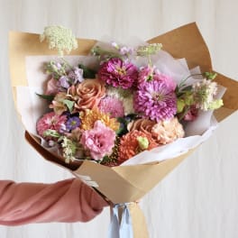 Hand-tied bouquet of pink, peach, and purple flowers wrapped in brown paper