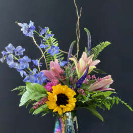 Tall mixed arrangement with pink lilies, blue delphinium, and a sunflower in an iridescent vase
