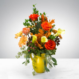 Tall autumn arrangement with orange roses, yellow calla lilies, gerbera daisies and orange leaves in a yellow vase.