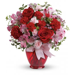 Red and pink roses, carnations, and alstroemeria in a red glass vase with a pink ribbon bow