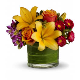 Low arrangement of yellow lilies, orange roses, purple alstroemeria, and red flowers in a clear glass vase