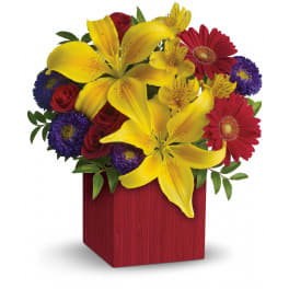 Bright arrangement of yellow lilies, red gerbera daisies, roses and purple asters in a red cube vase