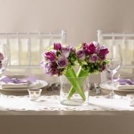 Three small arrangements of purple and lavender flowers in clear glass vases on a set dining table