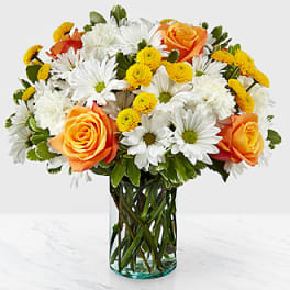 Mixed bouquet of orange roses, white daisies, and yellow poms in a clear glass cylinder vase