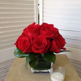 Red roses arranged in a square glass vase with a small candle nearby