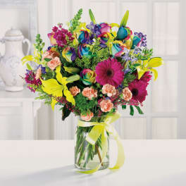 Colorful mixed bouquet with rainbow roses, yellow lilies, and pink gerberas in a glass vase with yellow bow