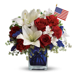Patriotic flower arrangement with red roses, white lilies, and blue accents in a blue glass vase with a small flag.