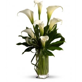 Tall arrangement of white calla lilies in a clear rectangular glass vase with looped green foliage.