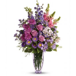 Tall arrangement of purple and pink roses, asters, snapdragons and mixed blooms in a slender lavender glass vase