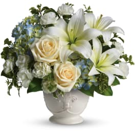 White lilies, cream roses, and blue hydrangeas arranged in a white ceramic pedestal vase