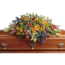 Colorful casket spray of mixed flowers including lilies, roses, and sunflowers on a wooden casket.