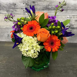 Bright mixed bouquet with orange gerberas, peach roses, and purple blooms in a green vase.