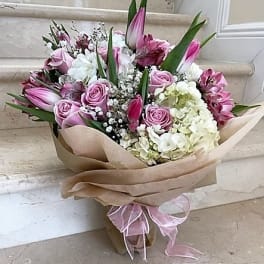 Hand-tied bouquet of pink roses, white hydrangeas, and pink lilies wrapped in kraft paper with a pink ribbon